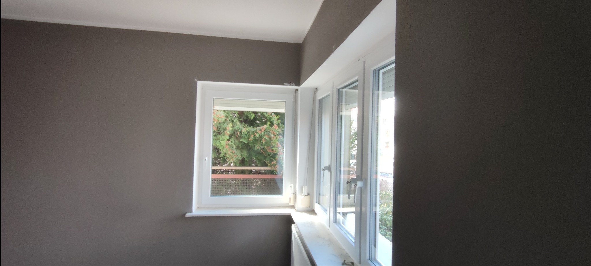 Ściana pomalowana na szaro, z białym oknem z widokiem na drzewa i balkon.