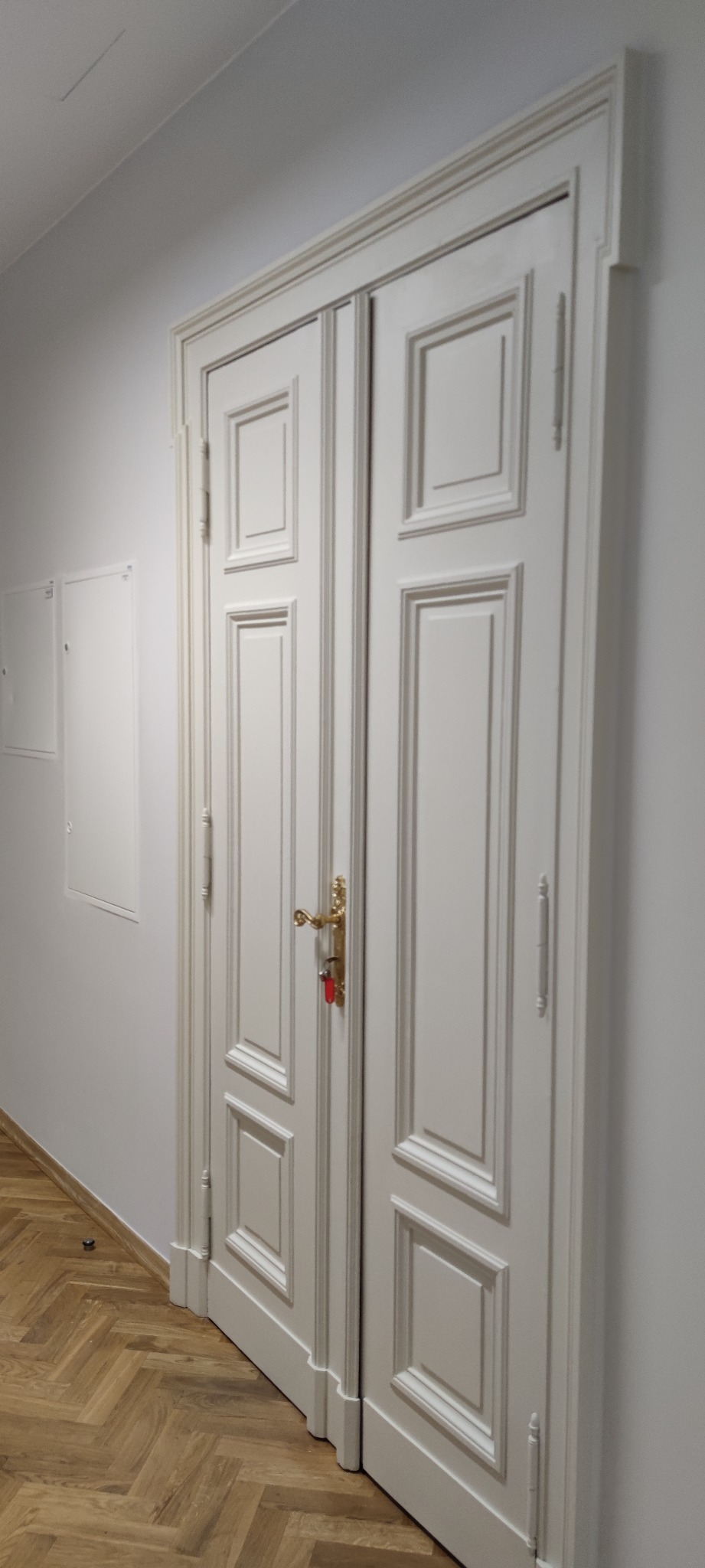 Białe, dwuskrzydłowe drzwi wewnętrzne z dekoracyjnymi panelami i złotą klamką z czerwonym breloczkiem, zamontowane w jasnym pomieszczeniu z parkietem ułożonym we wzór jodełki.