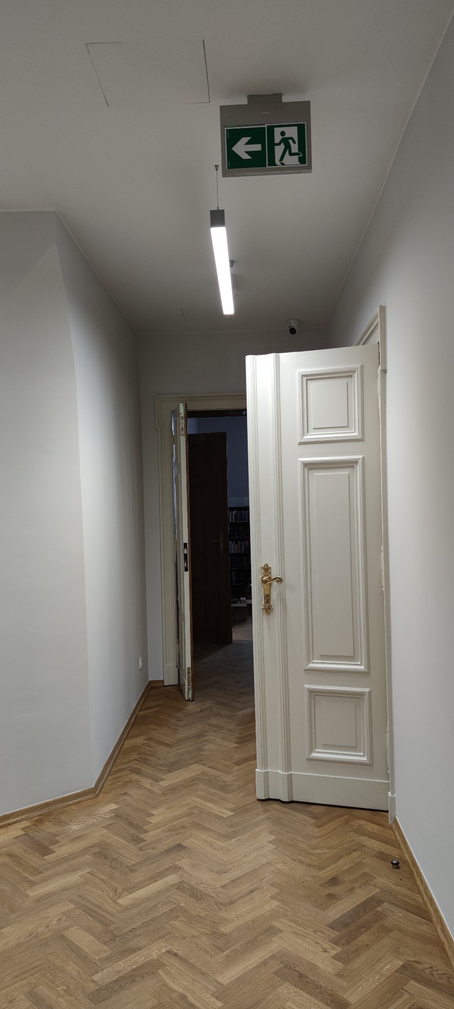 Korytarz z parkietem ułożonym w jodełkę, białe ściany, otwarte białe drzwi z ozdobną, złotą klamką, widoczny znak ewakuacyjny i oświetlenie sufitowe.