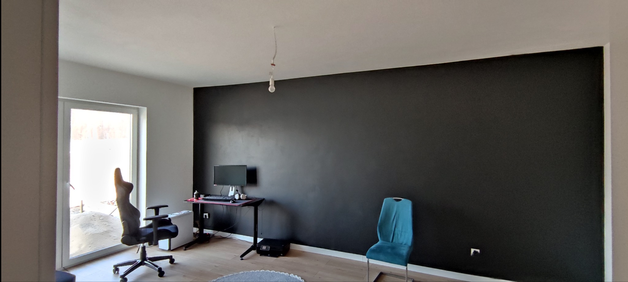 Pokój po malowaniu ścian na czarno, widoczny komputer, krzesło biurowe i turkusowe krzesło, surowa żarówka zwisająca z sufitu.