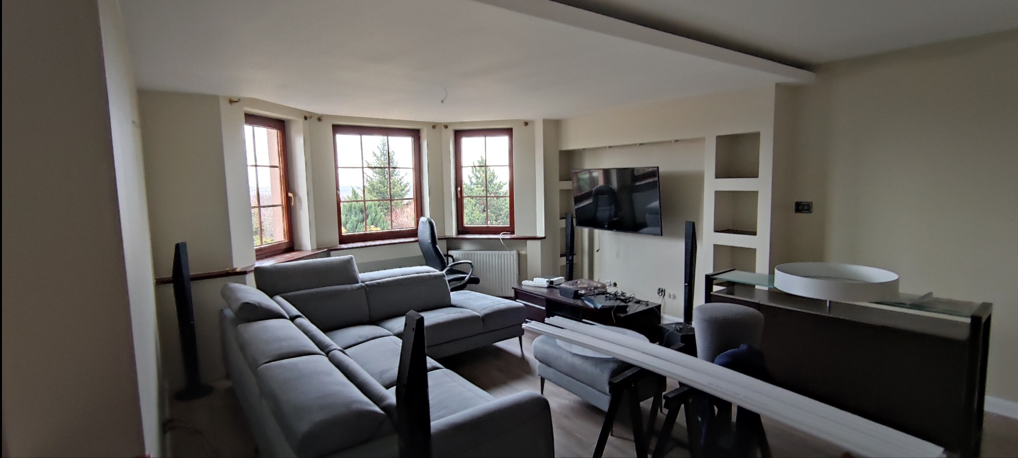 Salon w trakcie wykończenia: szara sofa narożna, trzy okna z widokiem na zieleń, telewizor na ścianie z półkami, listwy przypodłogowe na kozłach, drewniana podłoga.