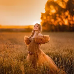Kobieta w złotej sukni, trzymająca się za brzuch, stoi na polu pszenicy o zachodzie słońca, z delikatnym logo fotografa w lewym dolnym rogu.