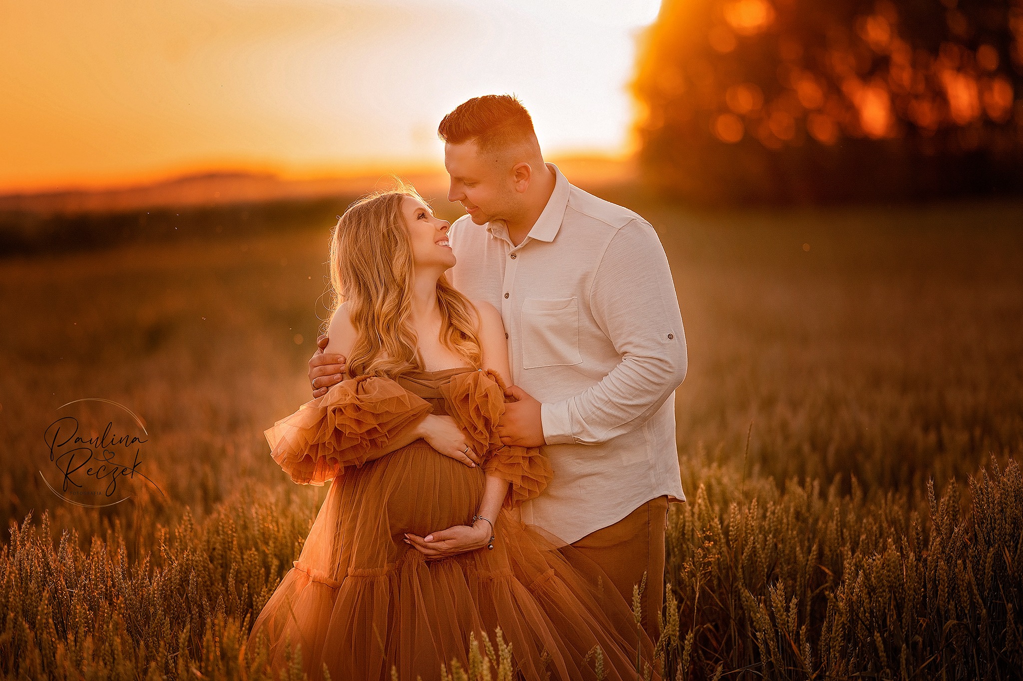 Przyszła mama w zwiewnej, brązowej sukni pozuje z partnerem na tle pola pszenicy o zachodzie słońca, oboje patrzą na siebie z uśmiechem.