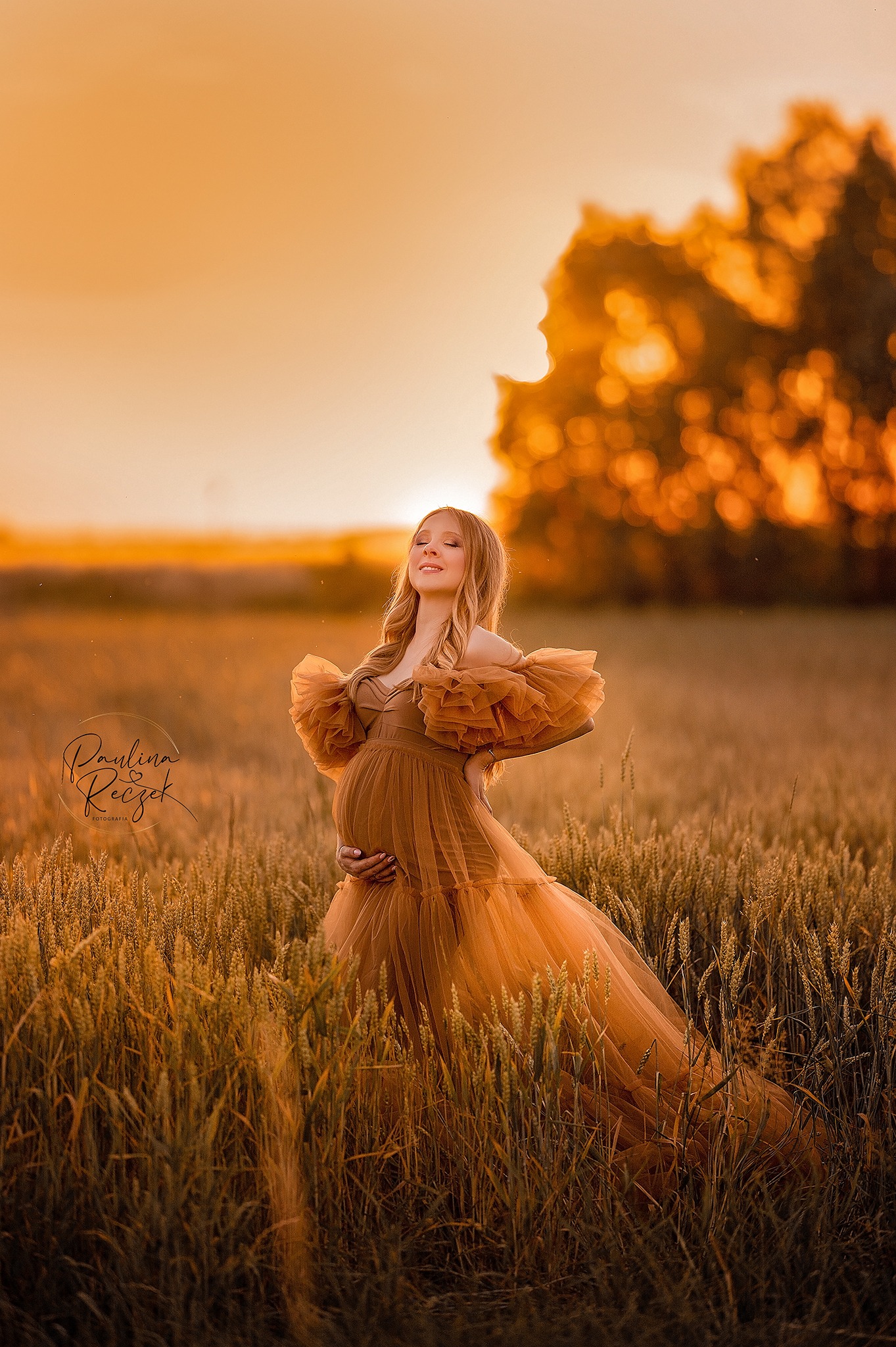 Kobieta w złotej sukni, trzymająca się za brzuch, stoi na polu pszenicy o zachodzie słońca, z delikatnym logo fotografa w lewym dolnym rogu.