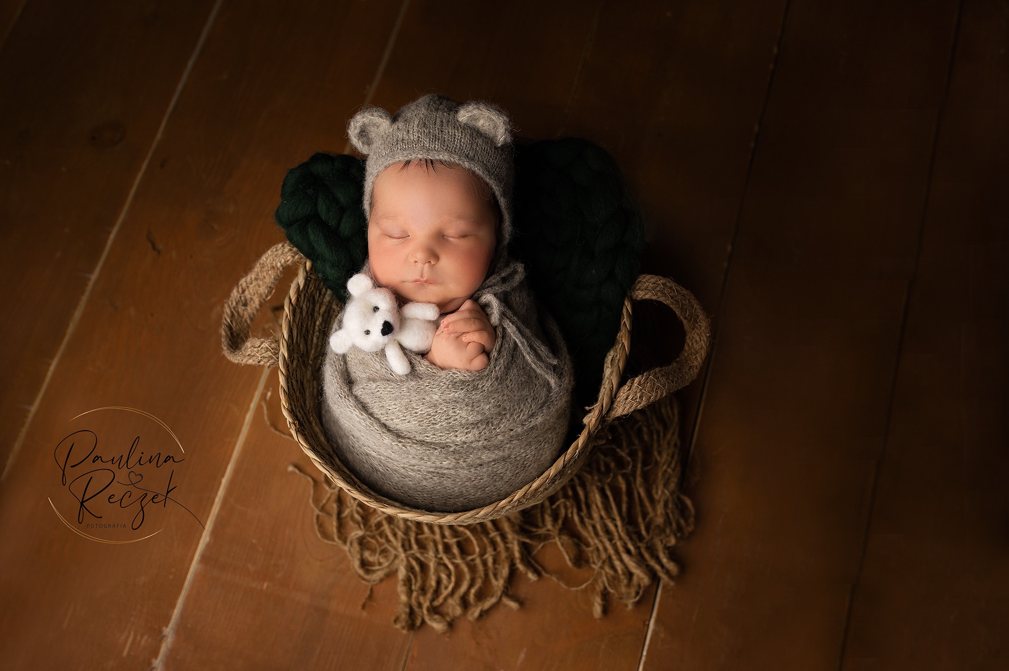 Śpiący noworodek w szarym kokonie i czapce z uszkami, trzymający pluszowego misia w wiklinowym koszu na drewnianej podłodze, z ciemnozielonym wypełnieniem pod głową.