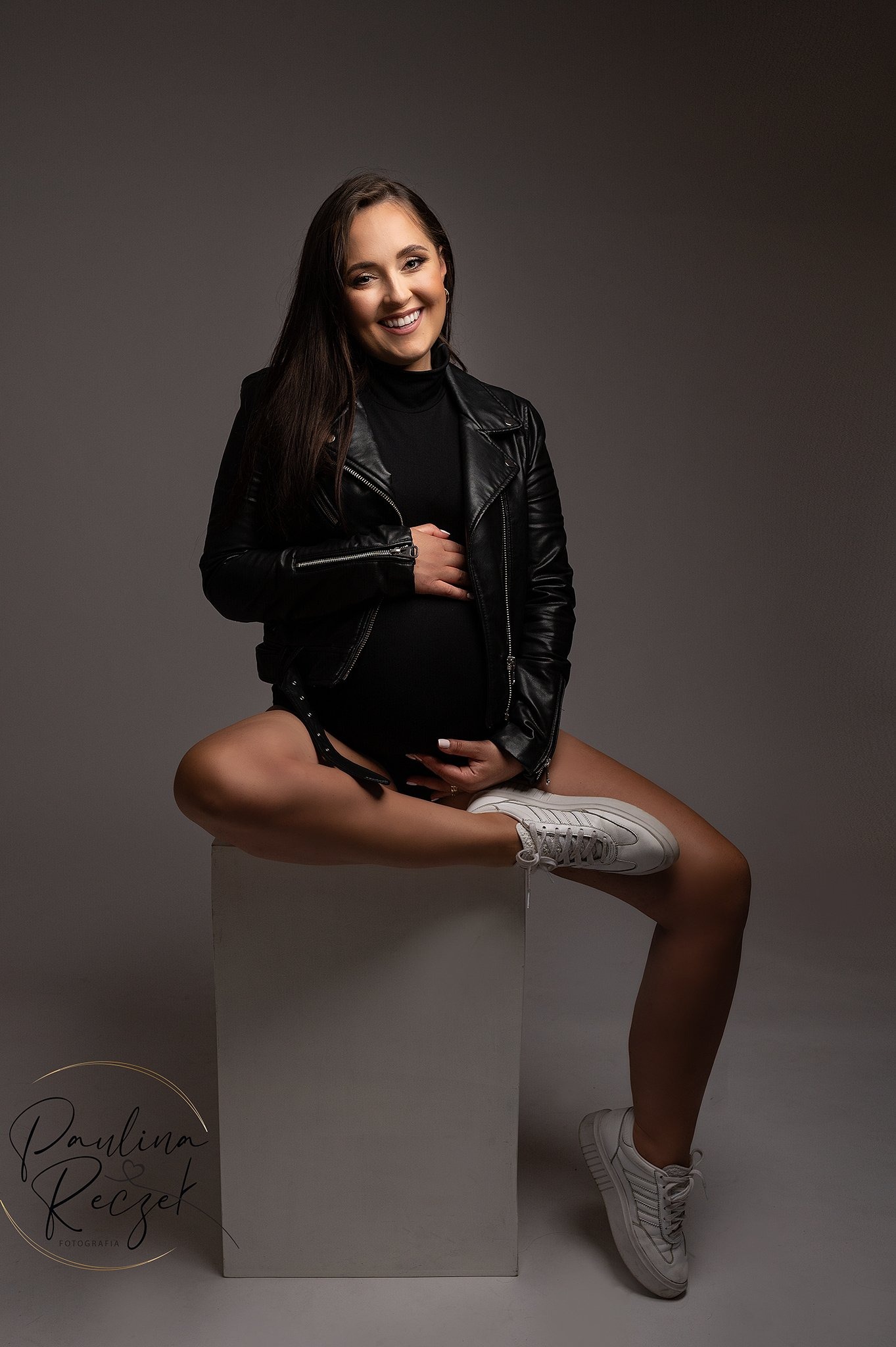 Uśmiechnięta, ciemnowłosa kobieta w ciąży, ubrana w czarny golf, skórzaną kurtkę i białe sneakersy, siedzi na białym sześcianie, trzymając dłonie na brzuchu na szarym tle.