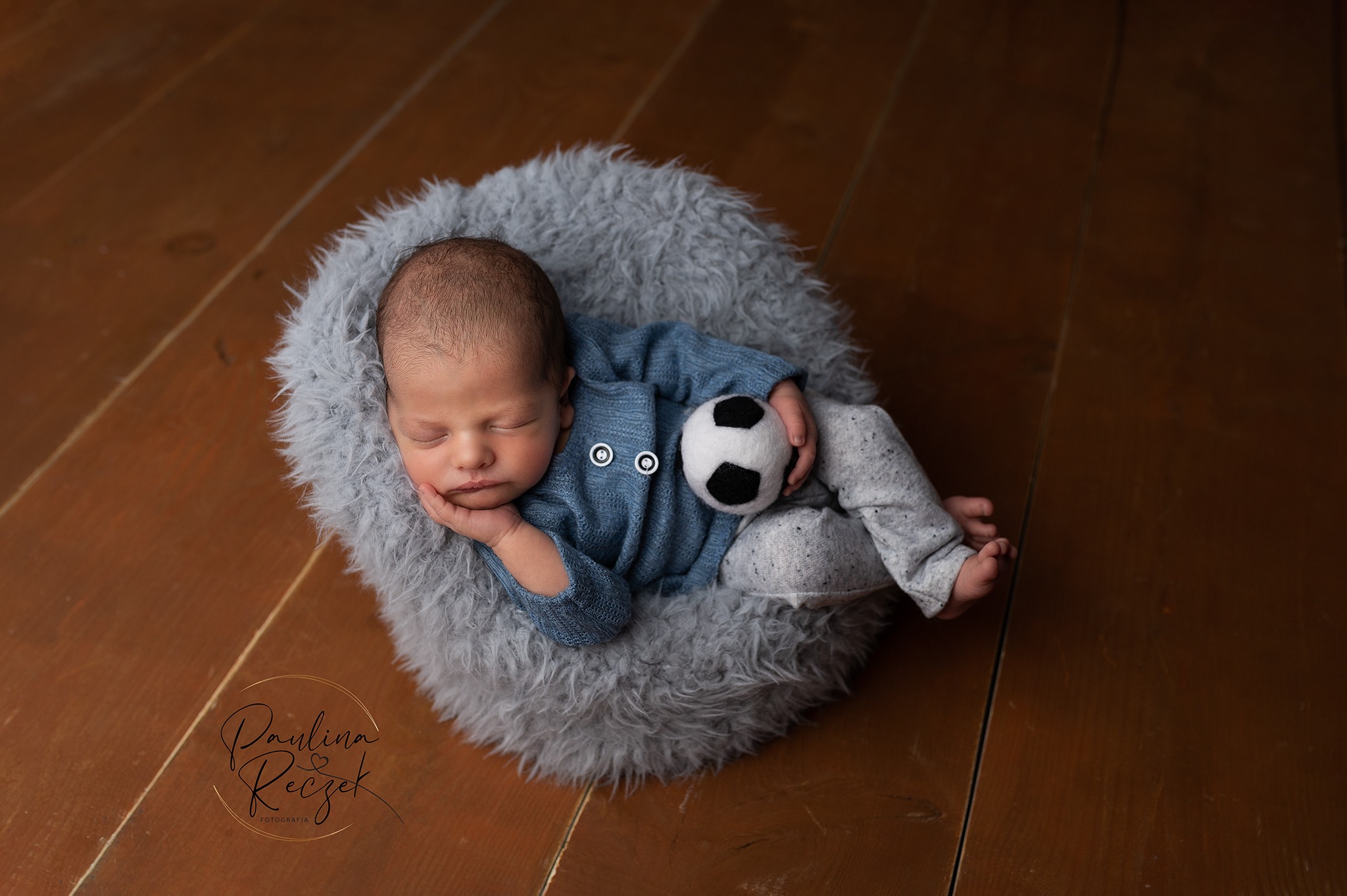 Noworodek śpiący na puszystym szarym podłożu w niebieskim sweterku, trzymający małą piłkę nożną, leżący na drewnianej podłodze. W rogu widoczne logo fotografa.