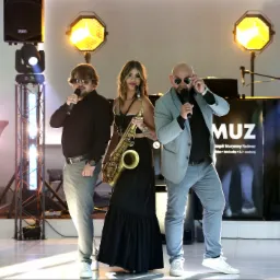 Zespół muzyczny Radmuz: wokalista w okularach przeciwsłonecznych z mikrofonem, saksofonistka z saksofonem i wokalista w marynarce poprawiający okulary, na tle sceny z oświetleniem i sprzętem...