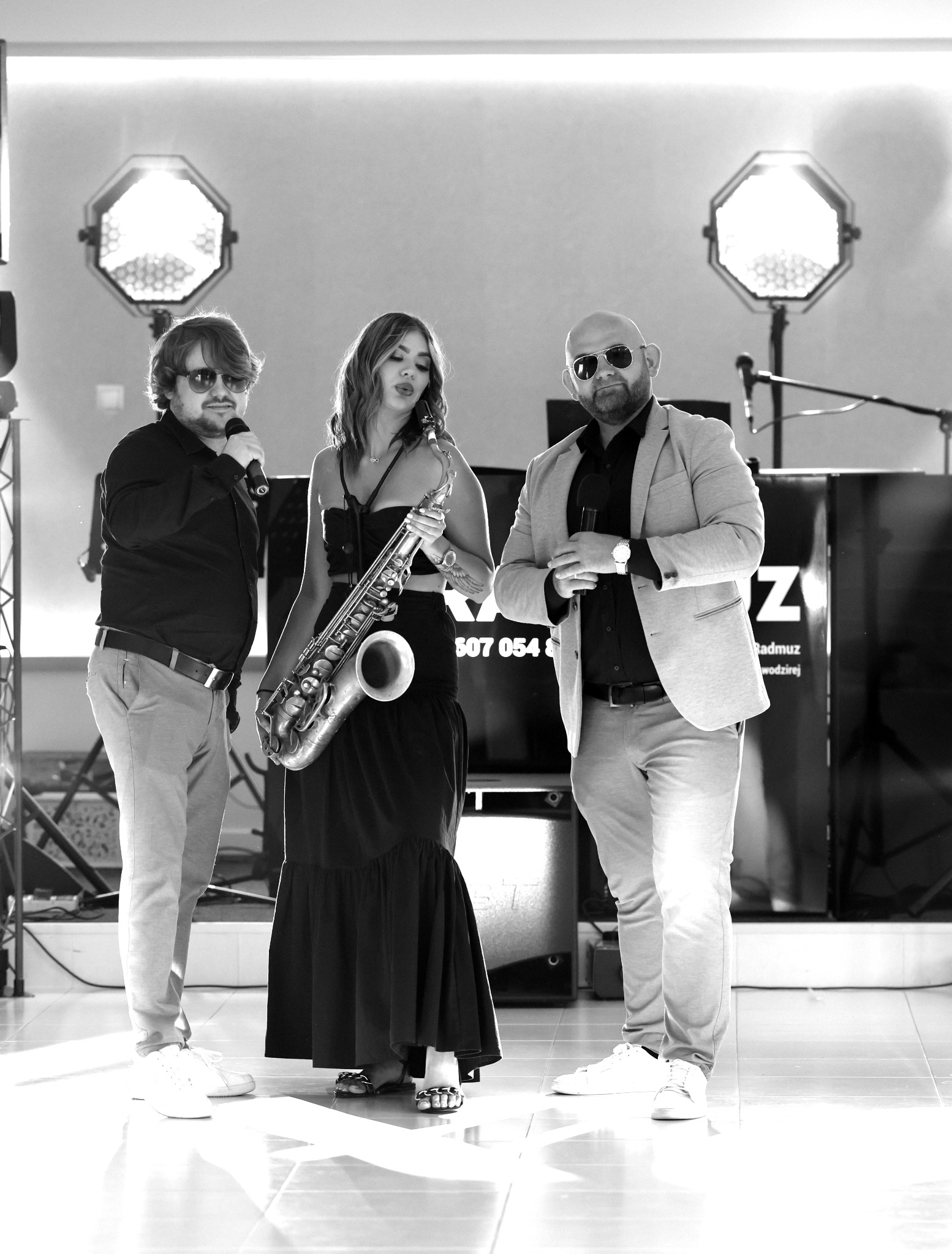 Czarno-białe zdjęcie zespołu muzycznego: wokalista z mikrofonem, saksofonistka i konferansjer w okularach słonecznych, oświetlenie sceniczne w tle.