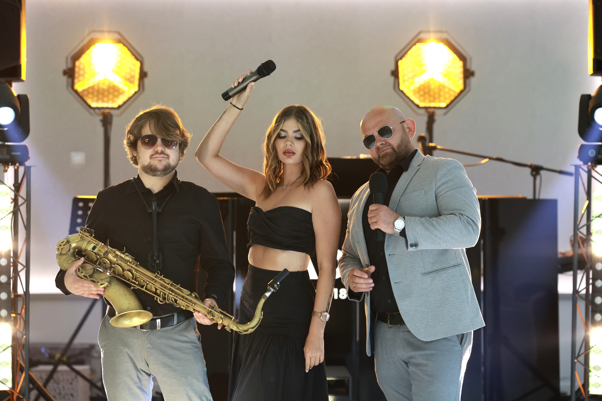Zespół muzyczny na scenie z saksofonem, wokalistką z mikrofonem i mężczyzną w marynarce z mikrofonem, oświetlenie sceniczne w tle.
