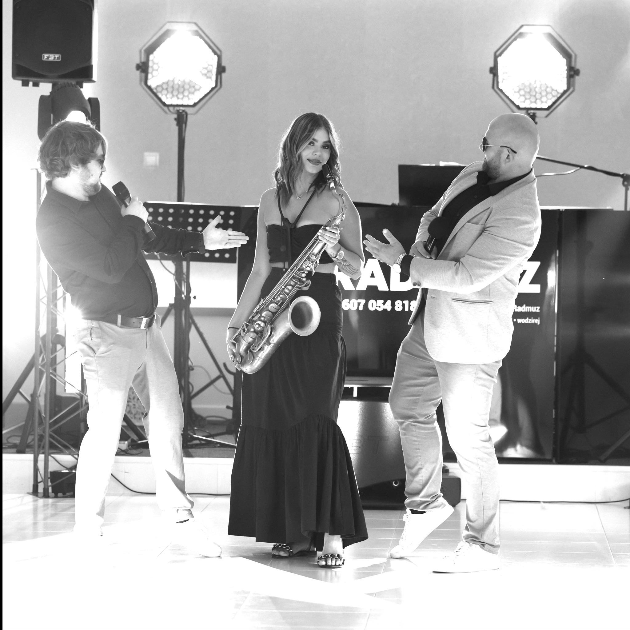 Czarno-białe zdjęcie zespołu muzycznego: wokalista z mikrofonem, saksofonistka w długiej sukni i mężczyzna w marynarce na scenie z oświetleniem estradowym.