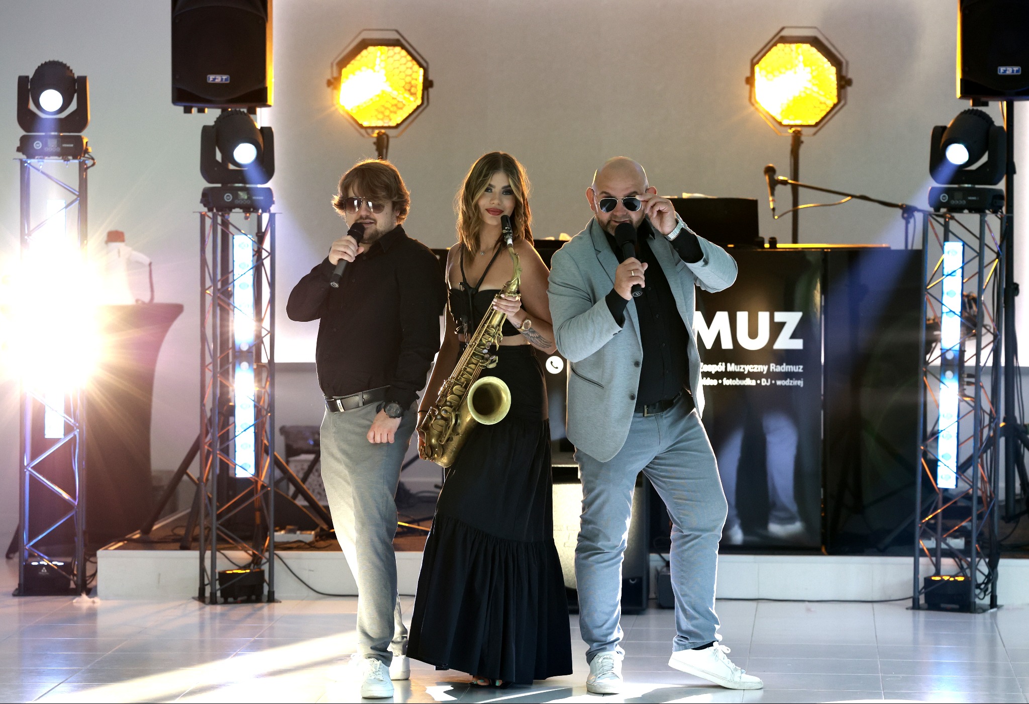 Zespół muzyczny Radmuz: wokalista w okularach przeciwsłonecznych z mikrofonem, saksofonistka z saksofonem i wokalista w marynarce poprawiający okulary, na tle sceny z oświetleniem i sprzętem...