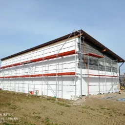 Dom jednorodzinny w trakcie prac związanych z ociepleniem elewacji styropianem, widoczne rusztowania, niezabezpieczona ściana z pustaków oraz stos palet w tle.