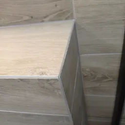 Szczegółowe ujęcie narożnika obłożonego płytkami imitującymi drewno, z widocznymi fugami i delikatnym wzorem słojów.