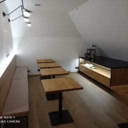 Wnętrze kawiarni na poddaszu z drewnianą podłogą, rzędem kwadratowych stolików z czarnymi nogami, wbudowaną ławą z jasną tapicerką wzdłuż ściany i kredensem z ekspresem do kawy.