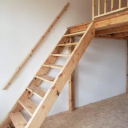 Surowe drewniane schody prowadzące na antresolę w Olsztynie, z widocznym fragmentem balustrady i niepomalowaną ścianą.