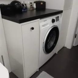 Biała szafka łazienkowa z czarnym blatem, obok pralka Samsung, na blacie kosmetyki i szkatułka, szara podłoga z ciemnymi fugami.