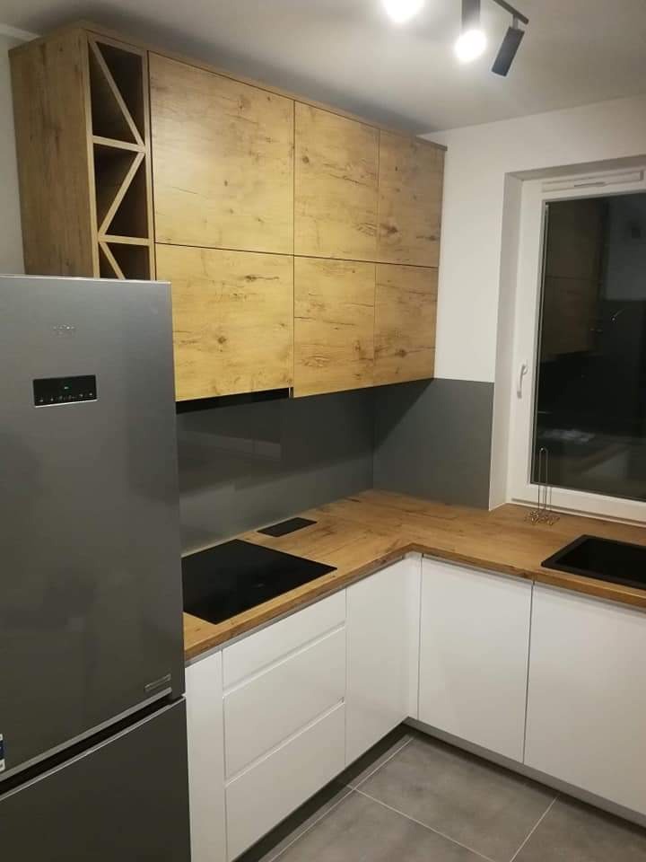 Nowoczesna kuchnia z białymi szafkami dolnymi, drewnopodobnym blatem i górnymi szafkami z drewnianym dekorem, obok srebrna lodówka i czarna płyta indukcyjna.