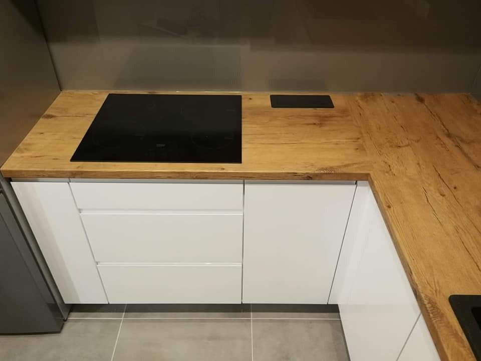 Kąt ujęcia na nowoczesną kuchnię z białymi szafkami i blatem z drewna o wyraźnym usłojeniu, z wbudowaną czarną płytą indukcyjną.