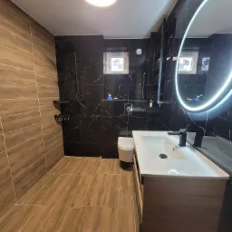 Nowoczesna łazienka z drewnopodobną podłogą i ścianą, kontrastującą z czarnymi płytkami imitującymi marmur na pozostałych ścianach, okrągłe lustro z podświetleniem LED, biała umywalka nablatowa...