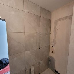 Patryk szafrański - Ściana łazienki w trakcie układania płytek imitujących marmur, widoczne krzyżyki dystansowe, fragment ściany przygotowanej pod dalsze prace, instalacja hydrauliczna.