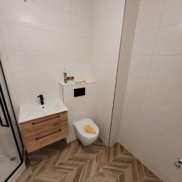 Patryk szafrański - Nowoczesna łazienka z drewnianą szafką pod umywalką, białymi płytkami na ścianach i podłogą ułożoną w jodełkę. Na spłuczce sedesu leży żółta ściereczka, a obok widoczny kartusz silikonu. Kabina...