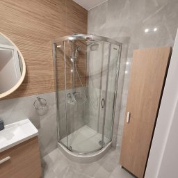 Patryk szafrański - Nowoczesna łazienka z kabiną prysznicową narożną, umywalką z szafką w kolorze drewna i płytkami imitującymi drewno na jednej ze ścian, a na pozostałych szare płytki imitujące marmur.