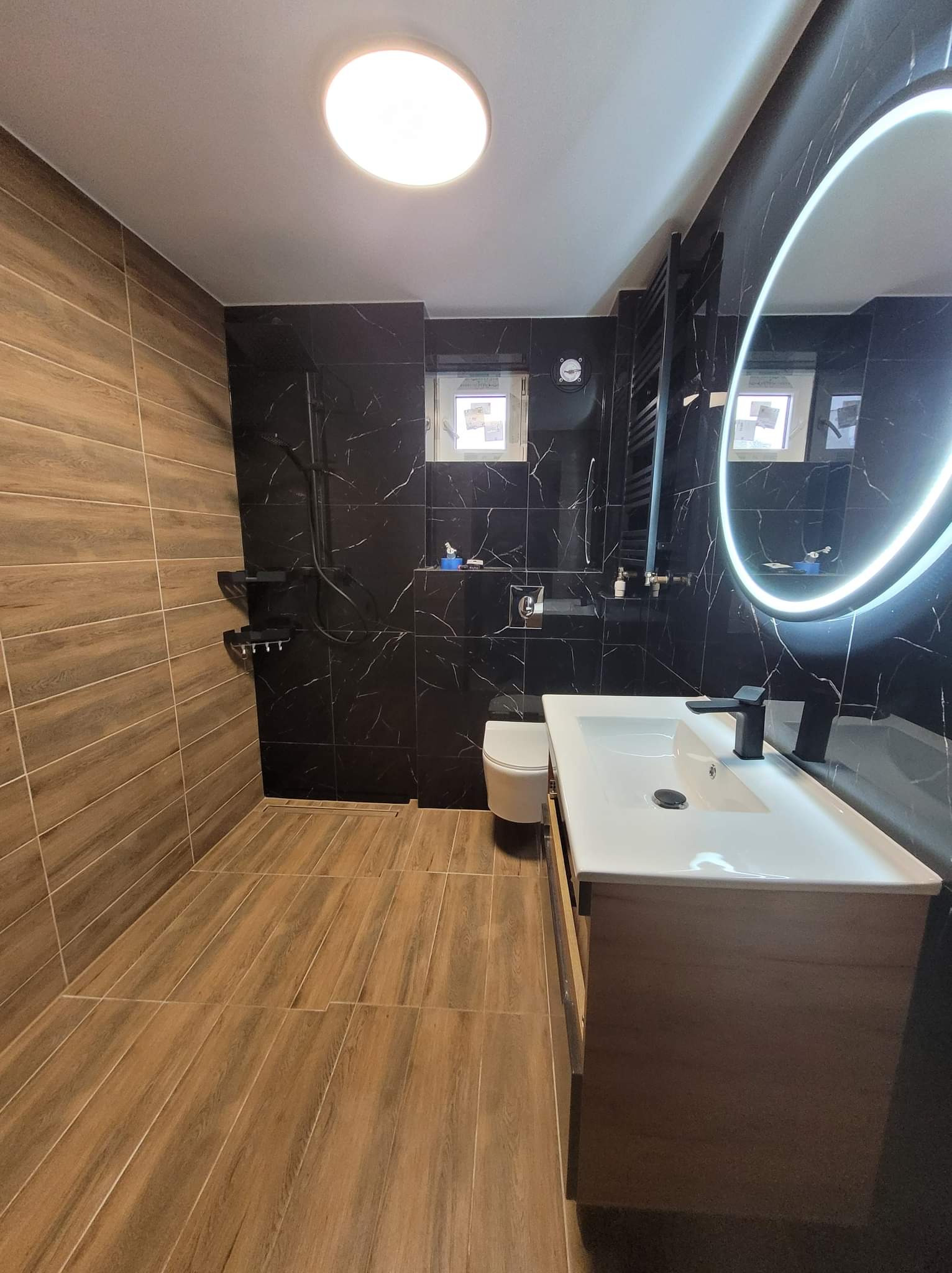 Nowoczesna łazienka z drewnopodobną podłogą i ścianą, kontrastującą z czarnymi płytkami imitującymi marmur na pozostałych ścianach, okrągłe lustro z podświetleniem LED, biała umywalka nablatowa...