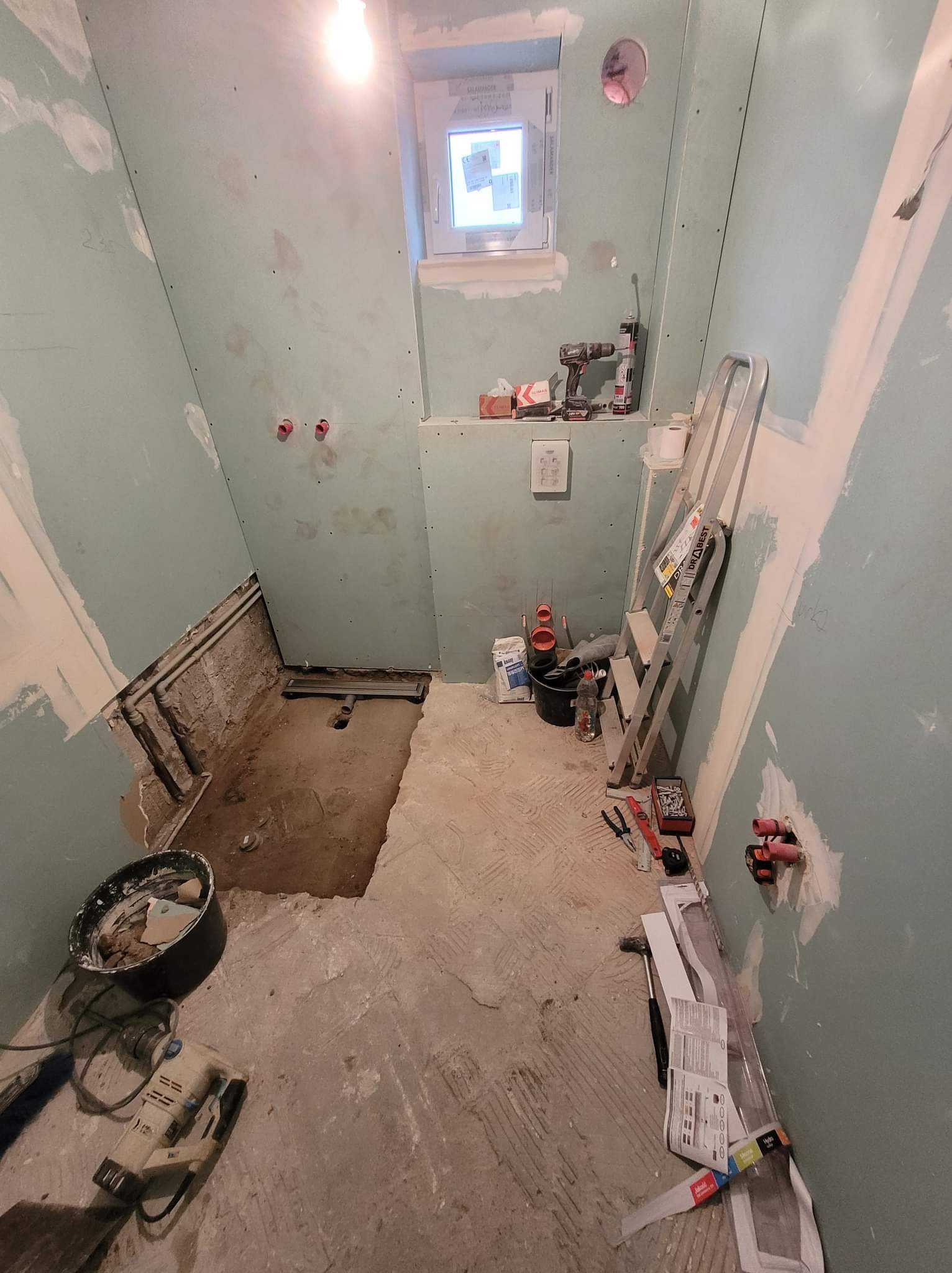 Łazienka w trakcie prac przygotowawczych: ściany pokryte płytami gipsowo-kartonowymi, widoczne instalacje hydrauliczne, narzędzia i drabina oparta o ścianę, surowy beton na podłodze.