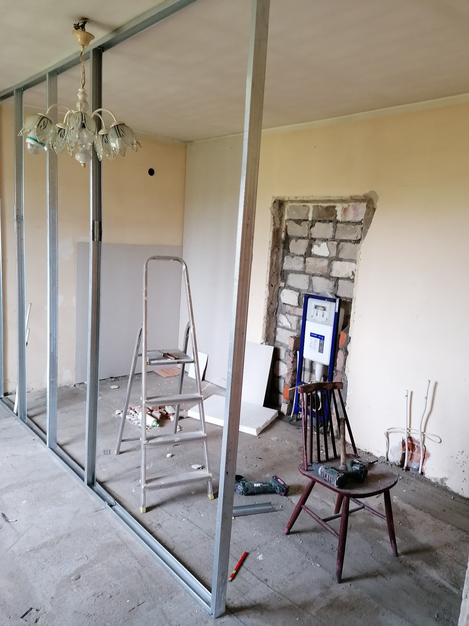 Remontowane wnętrze z metalową konstrukcją ścianki działowej, drabina aluminiowa, stara lampa wisząca i krzesło z wiertarką, widoczne instalacje hydrauliczne i elektryczne.