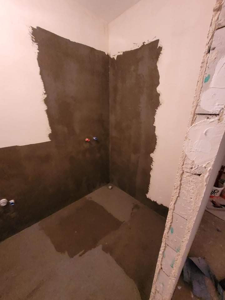 Niedokończona łazienka z częściowo nałożoną warstwą hydroizolacji na ścianach i mokrą podłogą, widoczne rury instalacji wodnej i fragment uszkodzonej ściany.