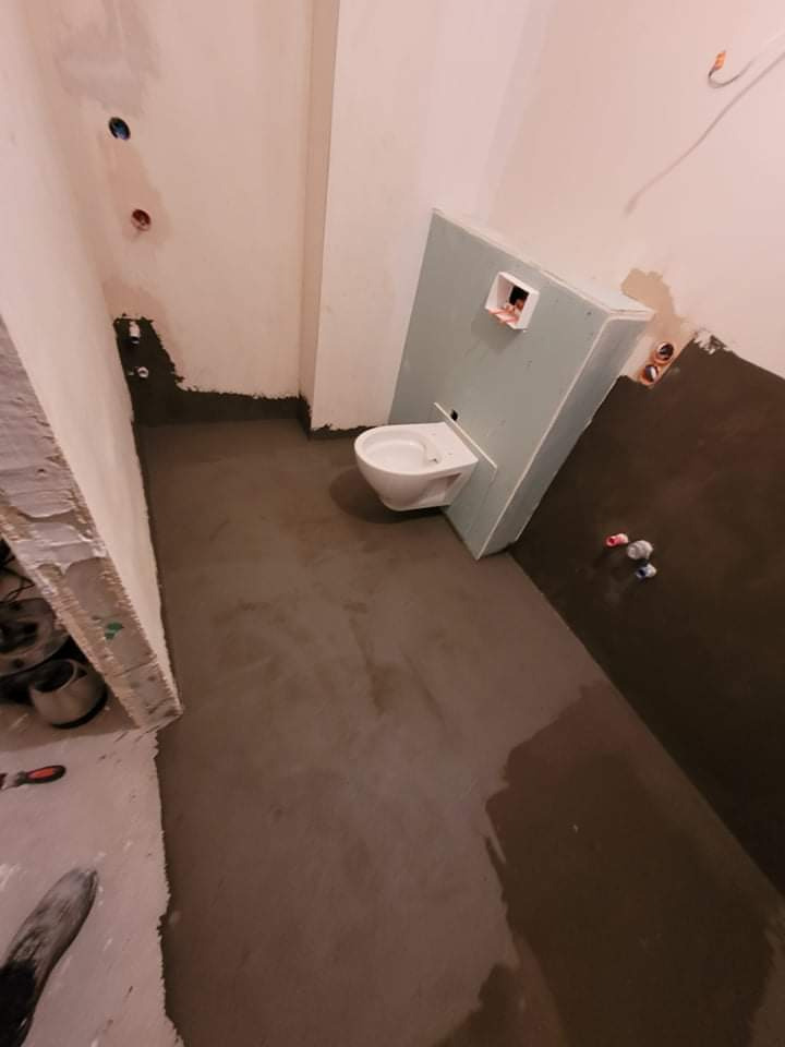 Świeżo wylana wylewka w łazience, widoczna podwieszana toaleta z zabudową z płyty gipsowej, przygotowane instalacje wodne i kanalizacyjne w ścianach, etap prac wykończeniowych.