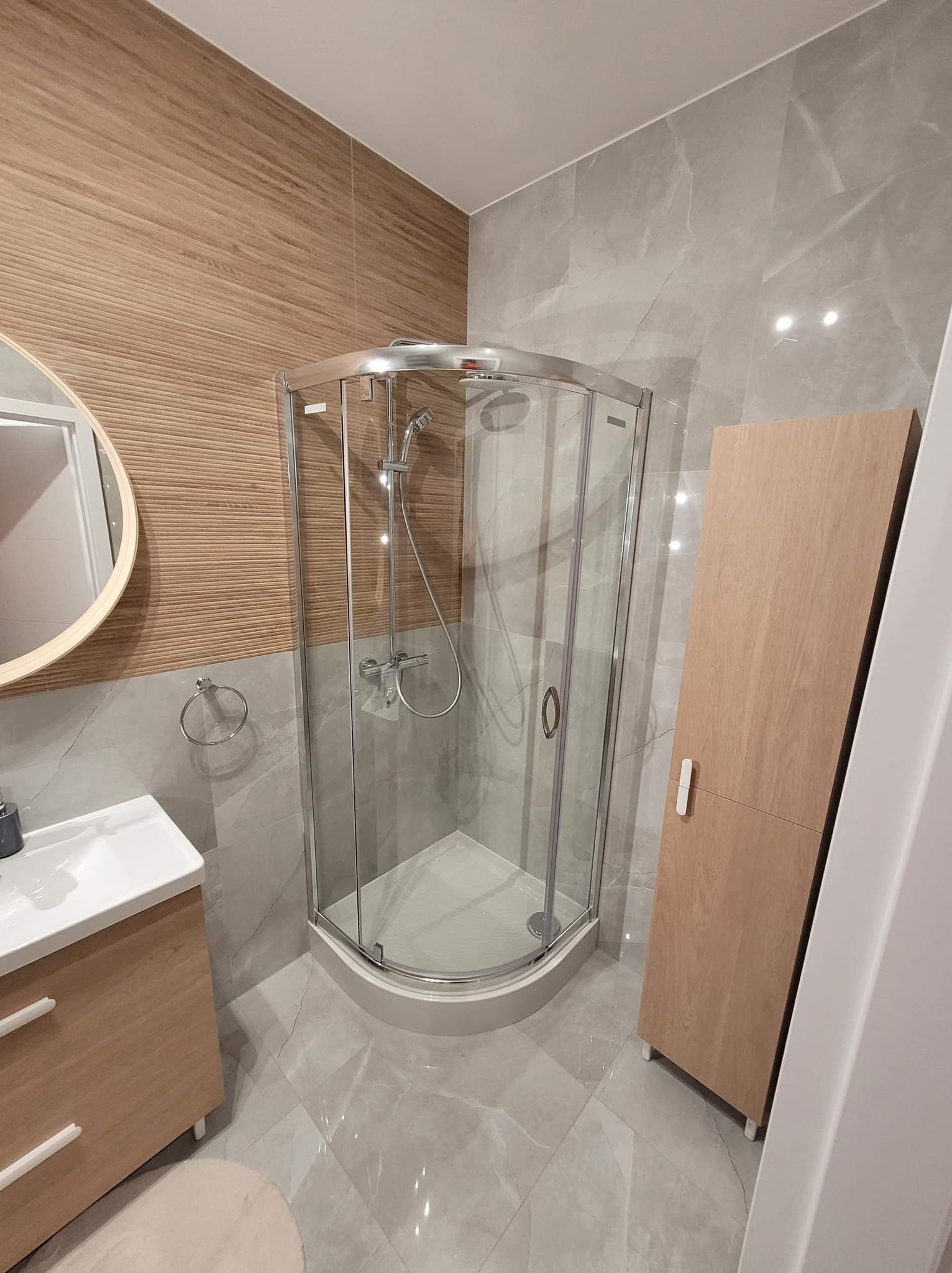 Nowoczesna łazienka z kabiną prysznicową narożną, umywalką z szafką w kolorze drewna i płytkami imitującymi drewno na jednej ze ścian, a na pozostałych szare płytki imitujące marmur.