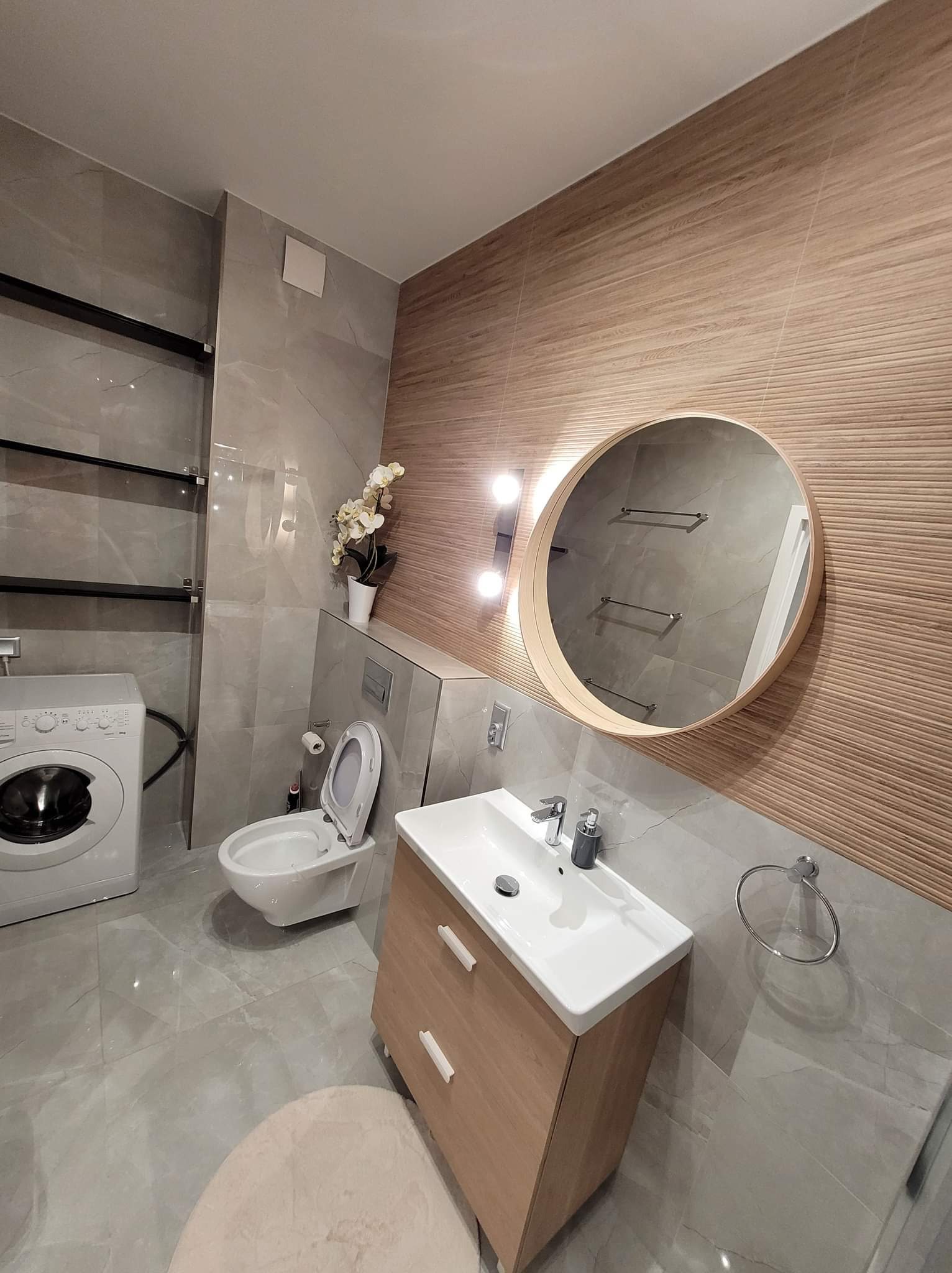 Nowoczesna łazienka z płytkami imitującymi drewno, okrągłym lustrem z podświetleniem, wiszącą toaletą, pralką i szarymi płytkami podłogowymi z widocznym okrągłym dywanikiem.