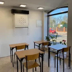 Sala z czterema biurkami i krzesłami, na ścianie tablica z diagramem. Jasne wnętrze, naturalne światło z okna. Gotowe miejsce do terapii grupowej lub zajęć.