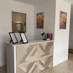 Pracownia psychologiczna mgr Małgorzata siwica - Recepcja z dekoracyjnym panelem z geometrycznym wzorem, dwie ramki z tekstami na blacie, obrazy kwiatów na ścianie i lampka biurkowa.