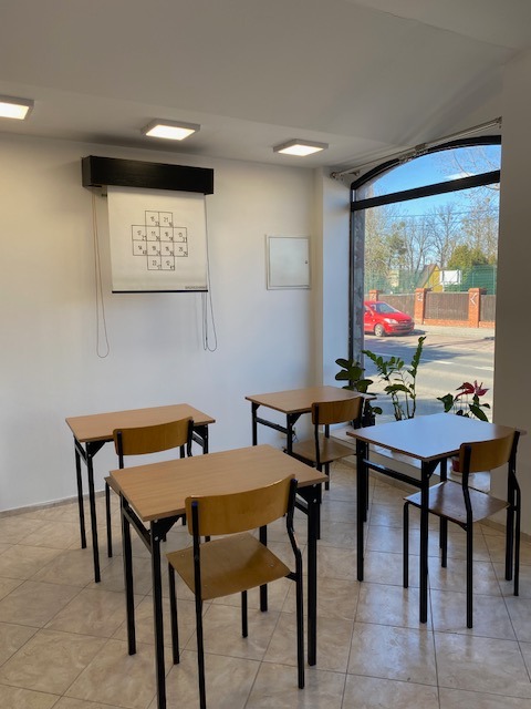 Sala z czterema biurkami i krzesłami, na ścianie tablica z diagramem. Jasne wnętrze, naturalne światło z okna. Gotowe miejsce do terapii grupowej lub zajęć.