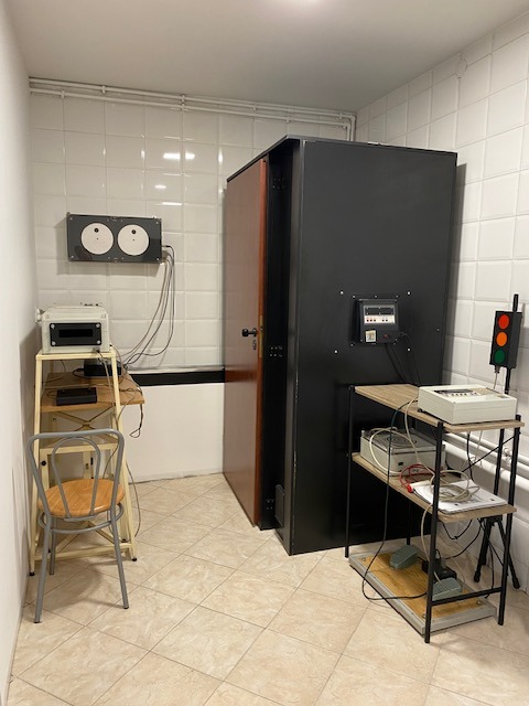Kabinowe stanowisko badawcze z aparaturą psychologiczną: kabina, stoliki z urządzeniami, lampa sygnałowa. Pomieszczenie wyłożone kafelkami.