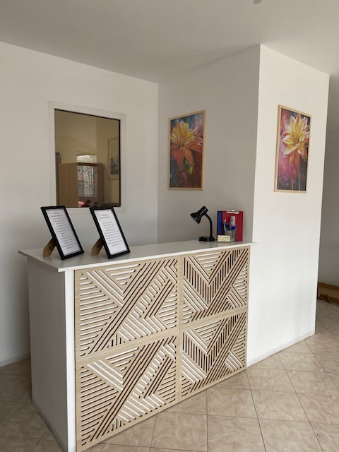 Recepcja z dekoracyjnym panelem z geometrycznym wzorem, dwie ramki z tekstami na blacie, obrazy kwiatów na ścianie i lampka biurkowa.