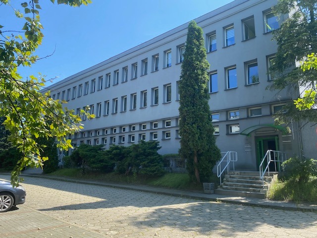 Szary budynek biurowy z zielonymi akcentami, widoczny z zewnątrz, z drzewem iglastym przed wejściem i fragmentem zaparkowanego samochodu po lewej.