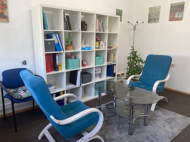 Jasne wnętrze gabinetu z dwoma turkusowymi fotelami, szklanym stolikiem i regałem z książkami. Na ścianie wiszą obrazy, a całość dopełnia roślina doniczkowa.