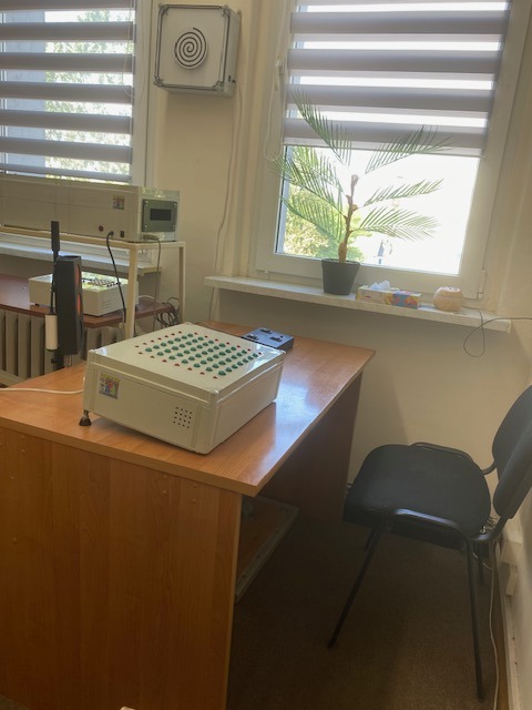 Gabinet z biurkiem, na którym stoi białe urządzenie z zielonymi diodami. W tle okno z roletami i roślina doniczkowa.