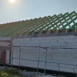 Budowa domu jednorodzinnego: ściany z betonu komórkowego, konstrukcja dachu z zielonych belek, widoczne rusztowania i drabina.