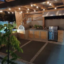 Zadaszona letnia kuchnia z piecem do pizzy, grillem gazowym i blatem roboczym, oświetlona girlandą żarówek, widok z zewnątrz.