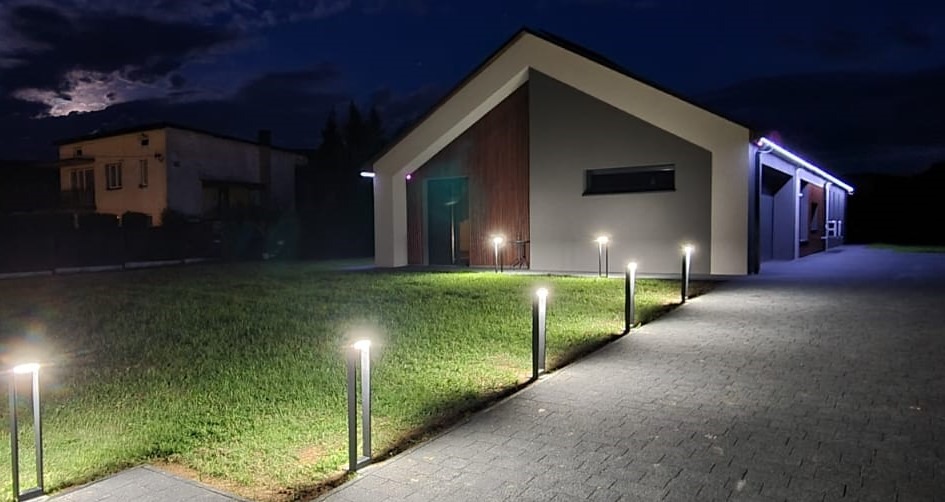 Nowoczesny dom jednorodzinny w Wadowicach oświetlony wieczorem lampami ogrodowymi wzdłuż ścieżki z kostki brukowej prowadzącej do wejścia.