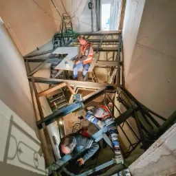 Trzech pracowników w kamizelkach odblaskowych montuje metalową konstrukcję pod sufitem w pomieszczeniu o surowych ścianach. Jeden siedzi na konstrukcji, drugi pracuje na niższym poziomie, a trzeci...