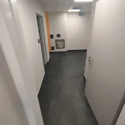 Korytarz biurowy z czarną podłogą, białymi ścianami i otwartymi drzwiami, widoczna wnęka z instalacją elektryczną i pomarańczowy akcent na ścianie.