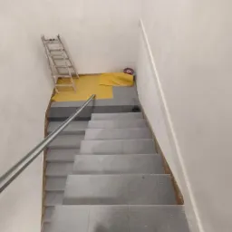 Klatka schodowa w trakcie remontu, zabezpieczona folią malarską, widoczna drabina oparta o ścianę i listwa przypodłogowa.