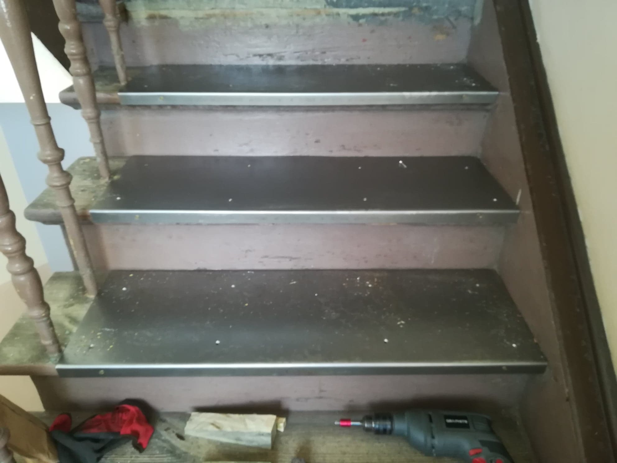 Schody drewniane z zamontowanymi metalowymi listwami na krawędziach stopni, widoczne ślady użytkowania i narzędzia pracy: wkrętarka, kawałki drewna i rękawice.