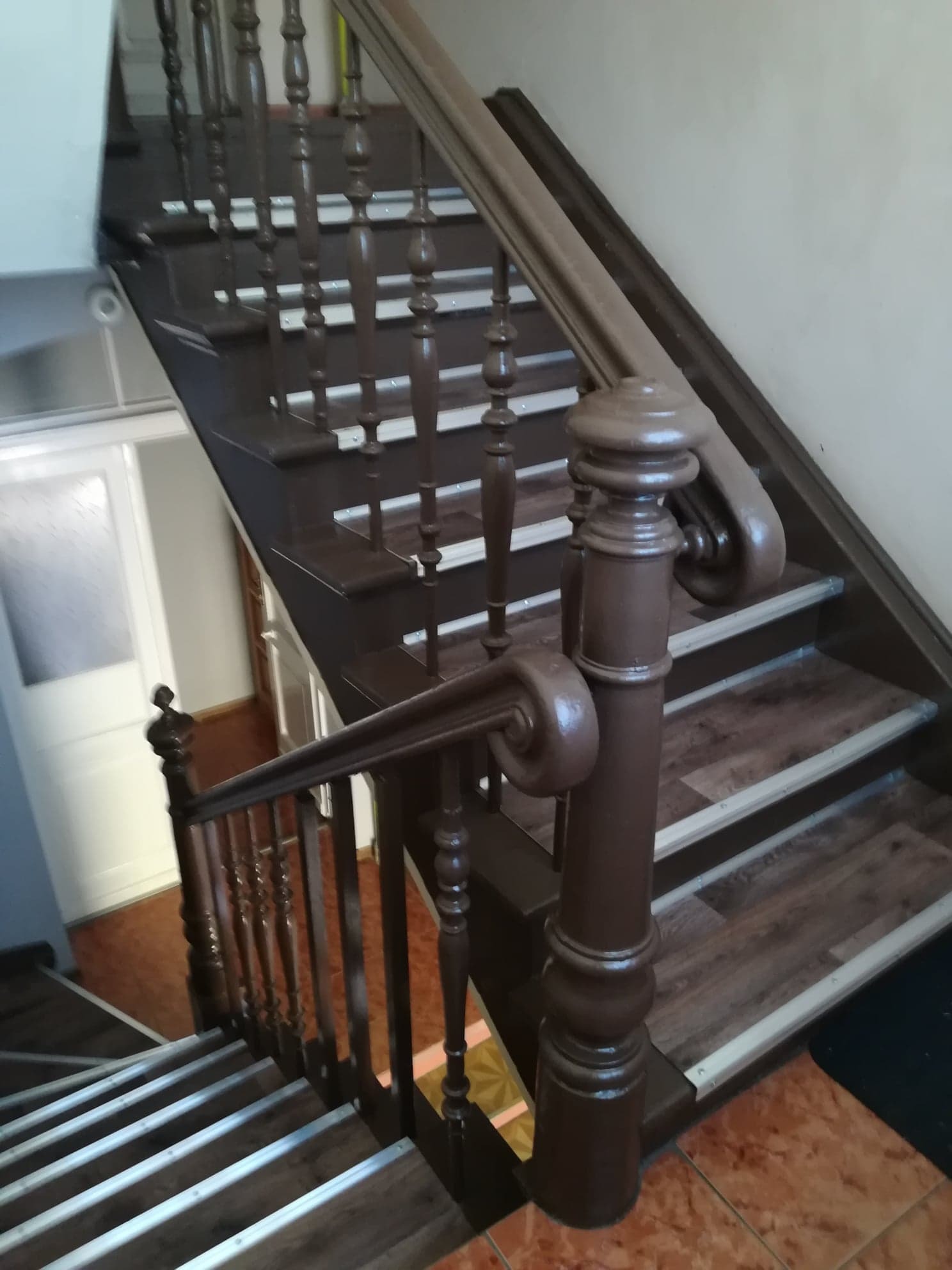 Klatka schodowa z drewnianymi schodami i brązową balustradą, widok z góry na dół, z widocznym wejściem do mieszkania w tle.